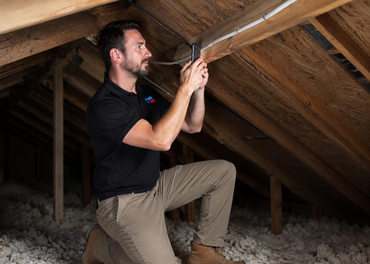 Roof Inspection Inside Attic | American Elite Roofing San Antonio Roofing professional kneeling inside a residential attic while taking inspection photos with a smartphone during a roof evaluation in San Antonio, Texas.